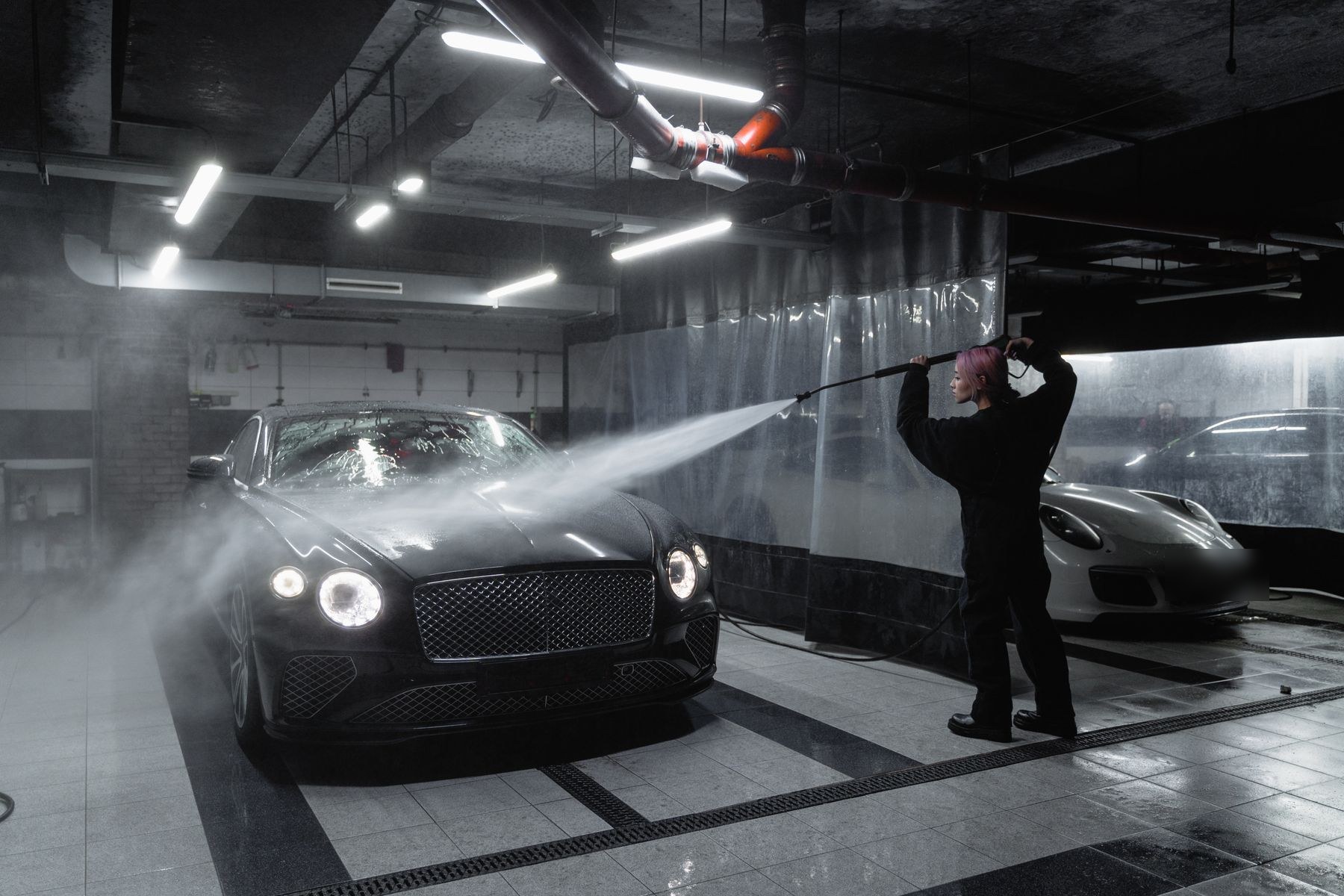Detailer rinsing a black luxury coupe in a dimly lit detailing bay
