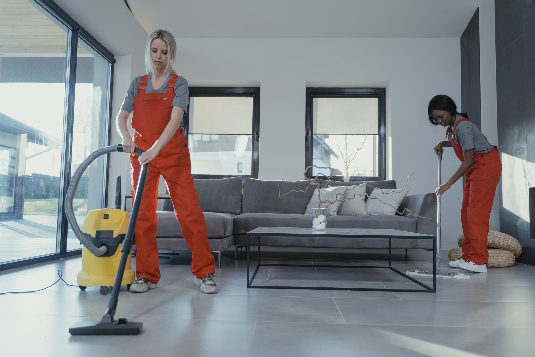Two cleaners in red coveralls vacuuming and mopping a bright living room